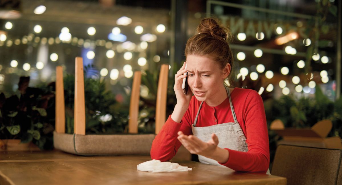Upset waitress sitting by table and talking to someone by smartphone at the end of working day