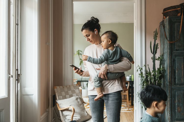 Woman wearing wireless in-ear headphones standing with baby boy while using smart phone at home