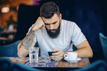 Man leaning on the table, holding smart phone and surfing on internet while sitting in cafe