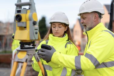 Two construction workers in bright safety gear surrounded by partially constructed steel frame building engage in conversation about project plans