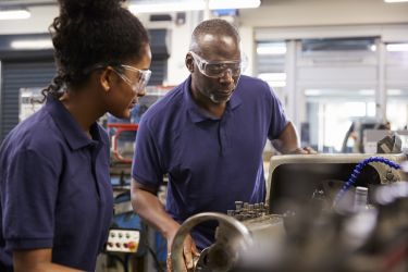 Older engineer showing female teenage apprentice how to use lathe 