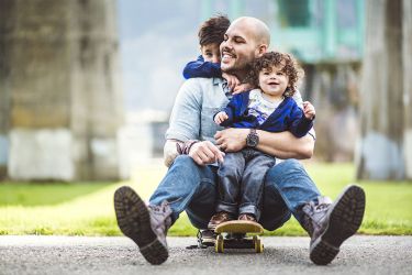 A father sits on a skateboard while hugging a child and being hugged by another child behind him