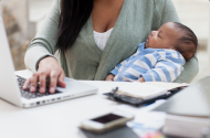 A woman works at a laptop while holding a baby