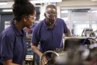 Older engineer showing female teenage apprentice how to use lathe 