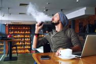 Young handsome hipster man with beard sitting in cafe with a cup of coffee, vaping and releases a cloud of vapor. Working at laptop and having a little break