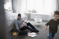 A man holding a baby attempts to work on a computer while another child runs across the bedroom