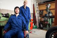 Two engineers in overalls; one young woman standing next to a young man in a wheelchair